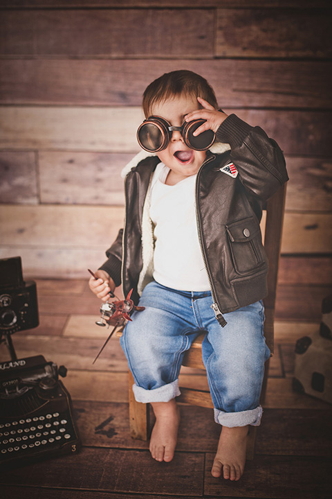 Foto de bebé con gafas y cazadora de aviador, con un avión en la mano y una máquina de escribir.