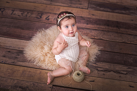 Fotografías de bebés, niña sentada en el suelo sobre una manta de pelo, jugando con un ovillo y mirando hacía el fotógrafo de bebé.