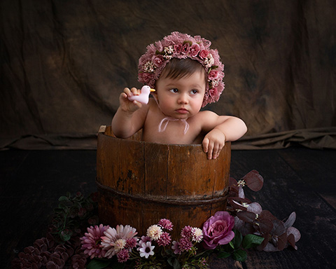 Niña dentro de un cubo con un gorro de flores jugando con un pato en su primera sesión de bebé.