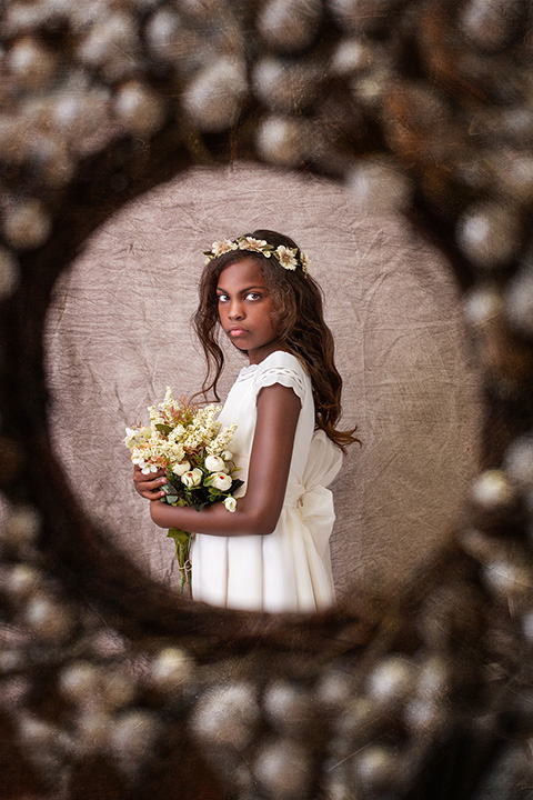 Niña morena con pelo largo y vestido blanco, sujeta unas flores entre sus manos y la rodea una corona de perlas blancas en su sesión de fotos de comunión en Madrid.