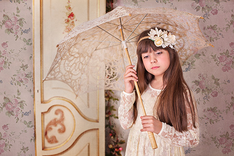 Fotógrafos de comunión en Madrid realizan sesión de una niña morena de pelo largo, con un tocado en la cabeza, lleva un vestido de encaje en color beige y está sujetando un paraguas entre sus manos, el fondo de la pared tiene flores rosas y hay una puerta antigua.