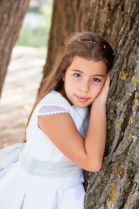 Medio plano de una niña con su mano y su cara apoyada sobre un árbol, lleva un vestido blanco y mira hacía la cámara en su sesión fotográfica de comunión.
