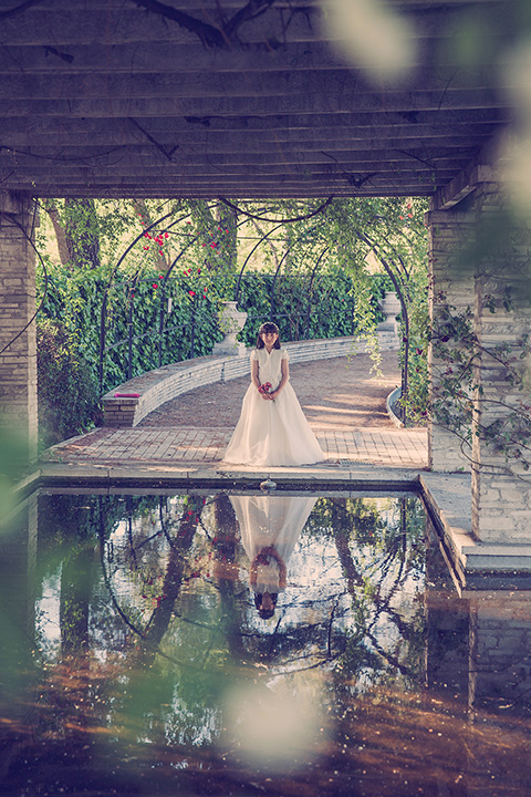 Plano general de una niña de pelo corto con un vestido blanco y unas flores entre sus manos, se está reflejando sobre una charca en su reportaje de comunión en exteriores. 
