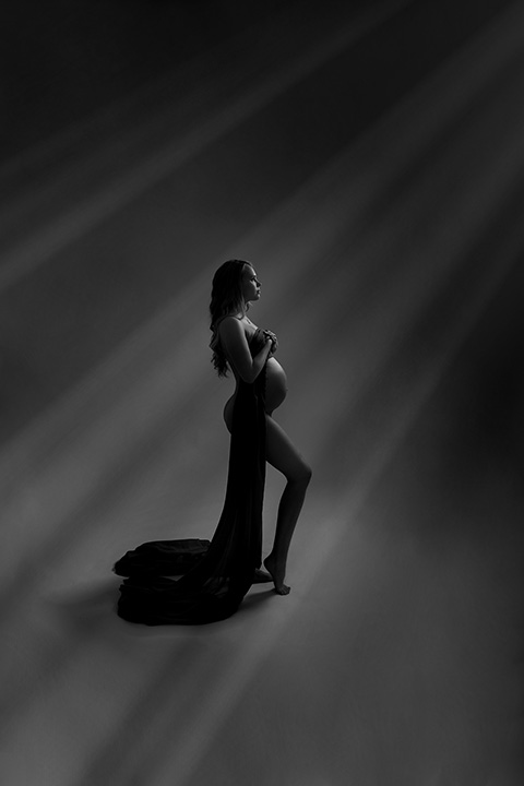 Mujer con una tela cubriendo su cuerpo, deja al descubierto su tripa y unos rayos muestran su contorno en una sesión artística de embarazo.