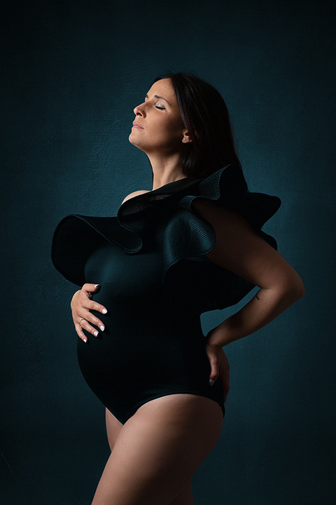 Fotografía de maternidad de medio cuerpo sobre un fondo azulado oscuro, la mujer tiene levantada su barbilla, ojos cerrados y su mano derecha toca su tripa.