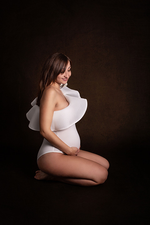 mujer sentada en el suelo, de rodillas, en fondo oscuro con body blanco con volantes marcando su figura en sesiones de fotos para embarazadas en Madrid.