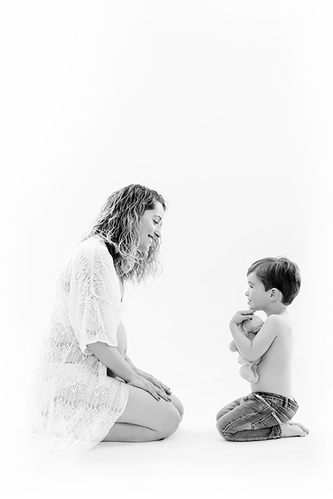 foto en blanco y negro de mujer sentada en el suelo frente a un niño que sujeta un conejo de peluche con sus manos pues están en una sesión fotográfica de embarazo con hijos.