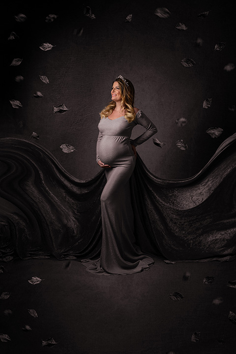 Imagen de mujer rubia con pelo largo, con vestido gris y con cola al vuelo, hojas volando a su alrededor pues está en una sesión fin art de maternidad.