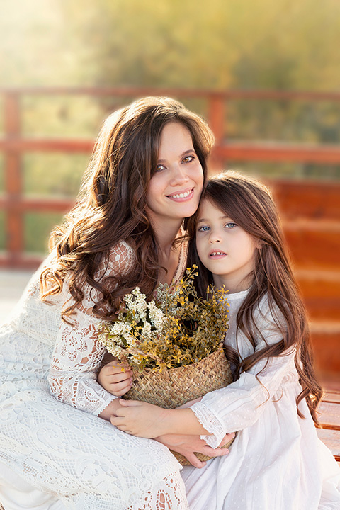 Hija con su madre en una sesión de fotos de familia en Madrid, están sentadas sobre un banco, tienen el pelo largo y llevan un vestido blanco, sujetan una cesta con flores entre sus manos.