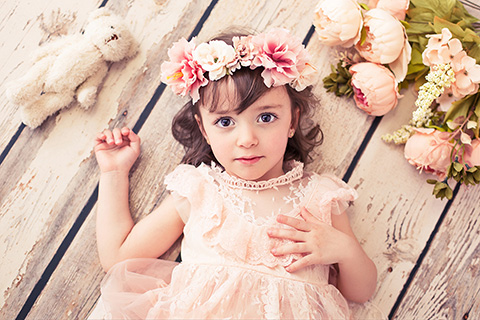 Niña tumbada en el suelo con una corona de flores rosas y un vestido a juego, a su lado tiene un peluche y un ramo, es una fotografía de moda infantil.