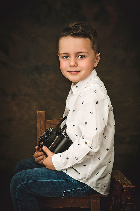 Niño con camisa blanca, sentado sobre una silla de madera, sujeta una cámara de fotos antigua entre sus manos realizada por un fotógrafo fine art en Madrid.