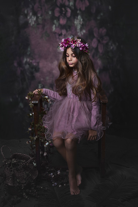 Niña morena de pelo muy largo con corona de flores en la cabeza en tonos púrpuras, con vestido a juego, está sentada en un banco imagen realizada por fotógrafo de moda infantil.