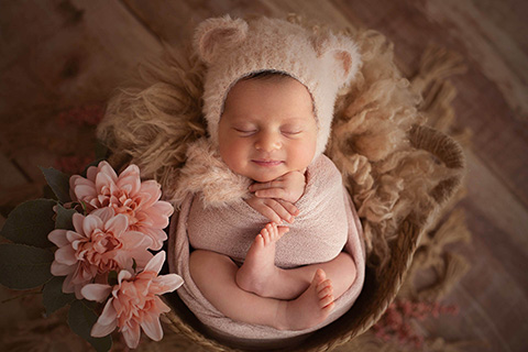 Niña con 15 días de vida, está dormida, metida en una cesta y tumbada boca arriba, está envuelta en una tela color salmón y lleva un gorro de pelo con forma de oso del mismo color.