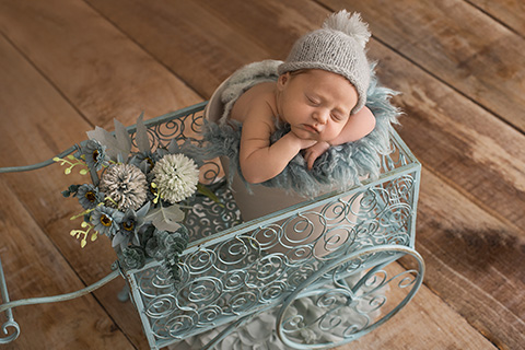Niña en su sesión para recién nacidos en estudio, está dormida dentro de un cubo de metal, sus manos y cabeza apoyan sobre una manta verde agua, lleva a juego un gorro de lana con pompón y está sobre un carro de hierro forjado del mismo color con flores.
