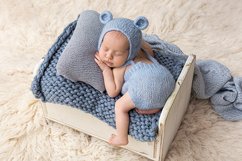 Bebé de tan solo 10 días, tumbado sobre una pequeña cama de madera blanca, lleva un conjunto de gorro de lana con orejas de oso y un peto en color azul, esta boca abajo, sus manos apoyan sobre una almohada y una de sus piernas cuelga hacia abajo, es una imagen realizada en un estudio de fotografía newborn.