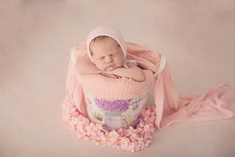 Niña dormida metida en un cubo de metal con dibujos de lavanda, tiene sus manos apoyadas sobre una manta rosa y su cabeza también apoya sobre ellas, lleva un gorro de lana rosa y alrededor del cubo hay muchas flores del mismo color, realizada por una fotógrafa newborn en Madrid.