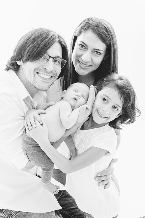 Padres e hijos posan en una sesión de fotos de recién nacido familiar, entre los tres sujetan al bebé que está dormido en sus brazos, visten camiseta blanca.
