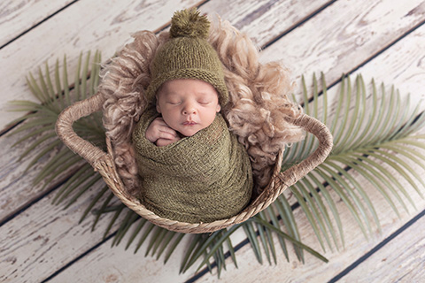 Bebé dormido en una cesta de paja, tiene el cuerpo en vuelta en una tela y sus manos asoma y se apoyan sobre su cara, lleva un gorro de lana con un pompón en color verte, alrededor tiene dos hojas de palmera realizada en una sesión de recién nacido original.